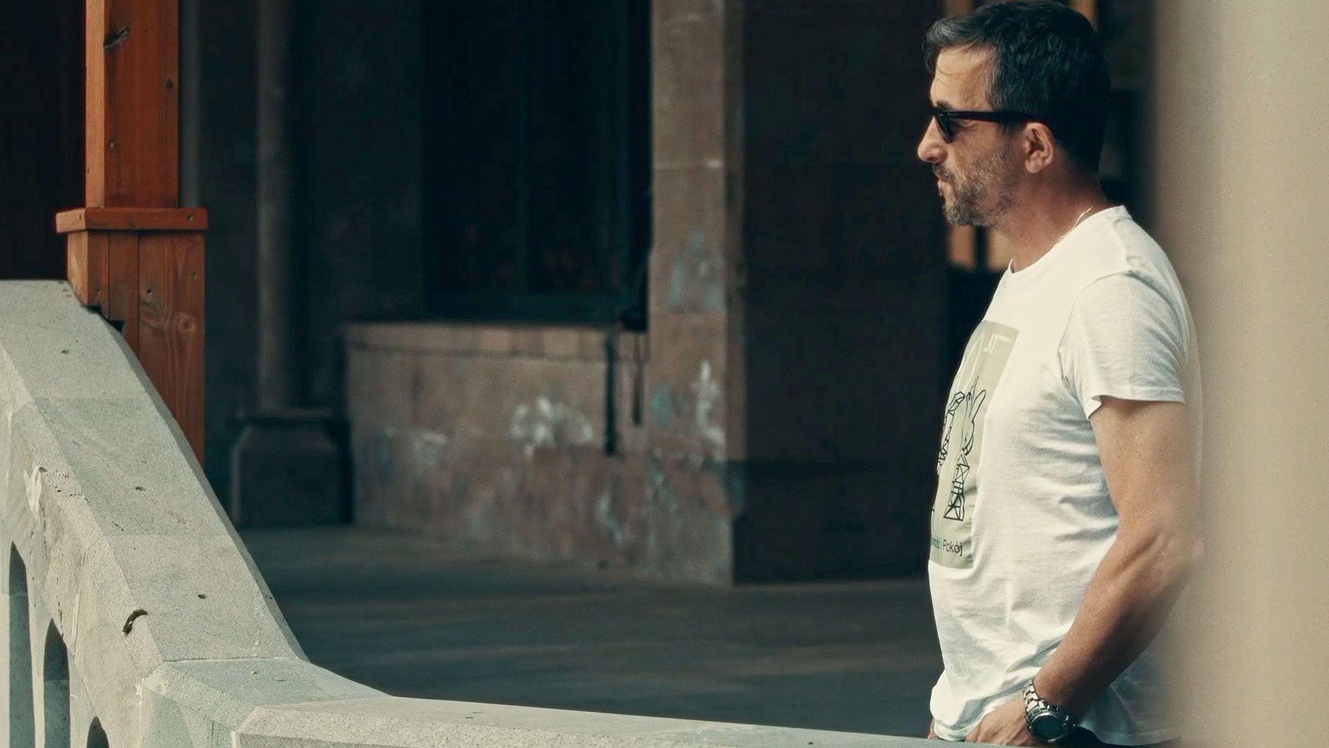 Marcin Mamon lleva una camiseta blanca y gafas oscuras. Está de pie junto a un muro de piedra, con las manos en los bolsillos. 