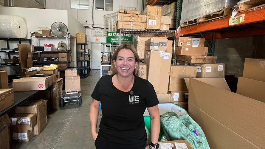 Una mujer vestida de negro sonríe en medio de un galpón lleno de cajas.