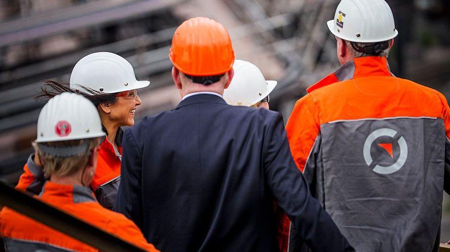 Varios trabajadores con mono naranja y en medio un ejecutivo vestido de traje.
