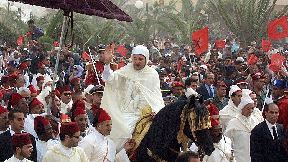 El rey Mohammed VI de Marruecos, ataviado con una larga túnica blanca tradicional, cabalga a caballo en medio de una gran multitud de personal de seguridad, oficiales militares, funcionarios y civiles, saludando a la multitud.