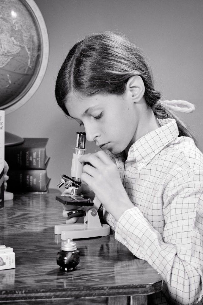 Años 60: una joven colegiala, absorta en sus estudios, sentada en el escritorio de su casa utilizando un microscopio para principiantes, rodeada de un globo terráqueo y libros de ciencia ficción.
