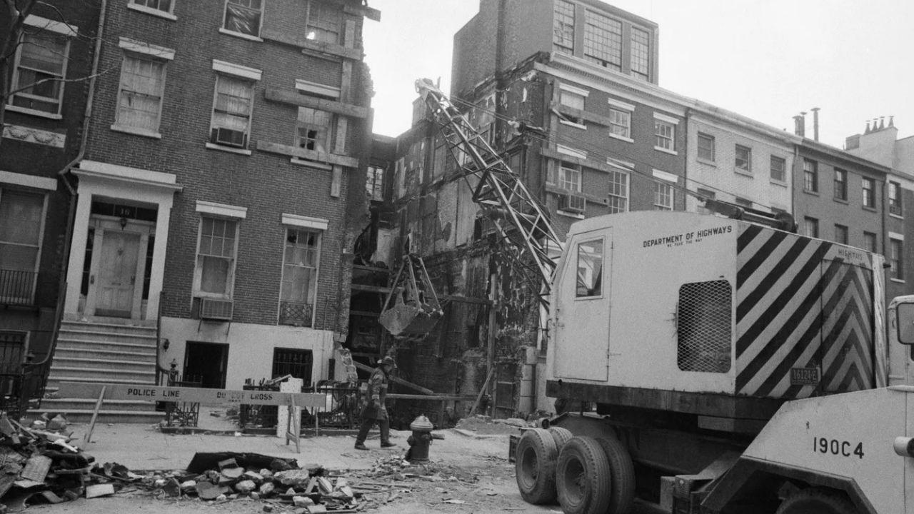 La fotografía en blanco y negro muestra una gran grúa móvil junto a una casa adosada de ladrillo gravemente dañada; la fachada y los pisos superiores del edificio parecen haberse derrumbado, dejando al descubierto las paredes interiores y los escombros; se puede ver a un trabajador cerca de los restos y una barrera con las palabras "línea policial" acordonó la zona.