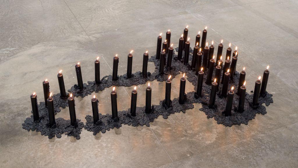 47 velas negras altas encendidas y dispuestas de manera que formen una silueta abstracta en el suelo de mármol