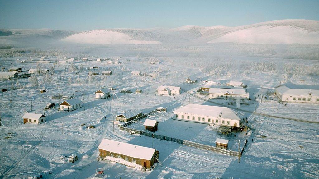 Vista aérea de un pueblo con varias construcciones bajas dispersas en una llanura nevada, con montañas cubiertas de nieve al fondo.