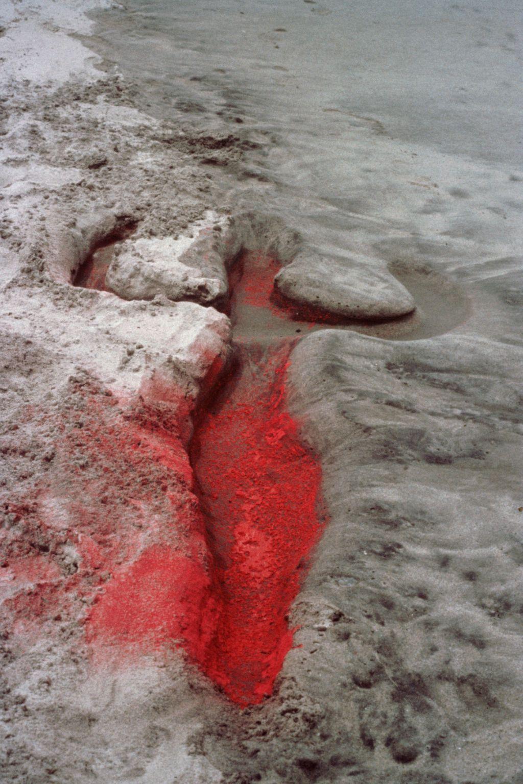 Una silueta femenina, tendida sobre la arena, aparece dibujada en el suelo con pigmento rojo. Tiene la cabeza redondeada, los brazos levantados y flexionados y las caderas anchas. 