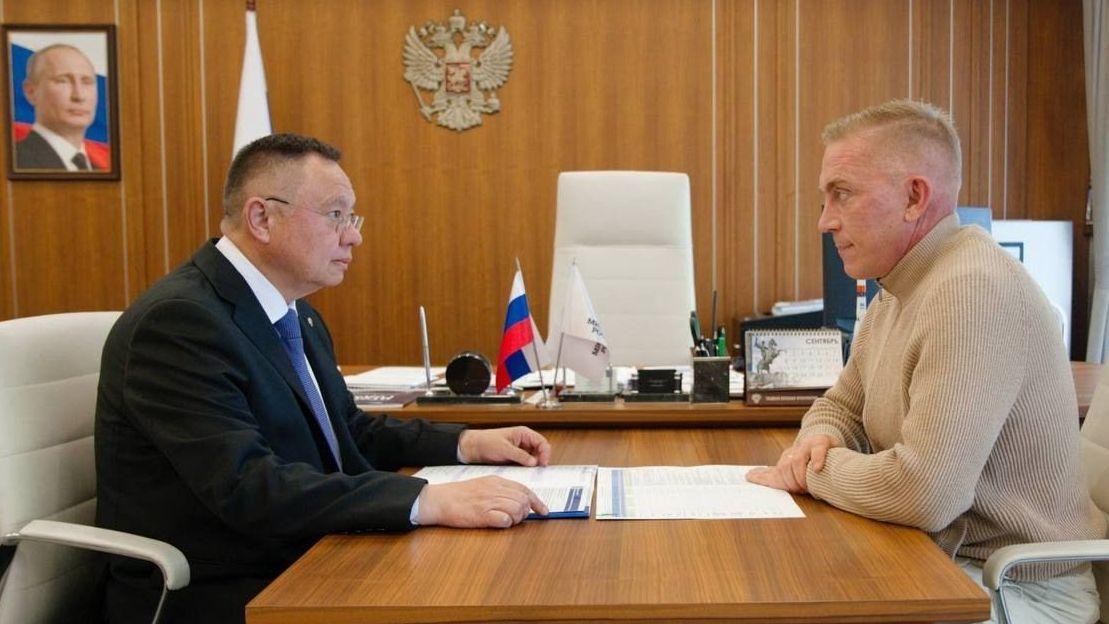 El ministro de Construcción, Irek Faizullin, con gafas, traje oscuro, camisa blanca y corbata azul, y el promotor inmobiliario del Grupo TSI, Alexei Fomenko, con jersey y pantalón color crema, están sentados en sillas del mismo color, frente a un escritorio lleno de documentos, en un despacho revestido de paneles con el escudo de armas ruso y un retrato del presidente Vladimir Putin, con traje marrón oscuro, camisa blanca y corbata a juego, frente a la bandera, en la pared.