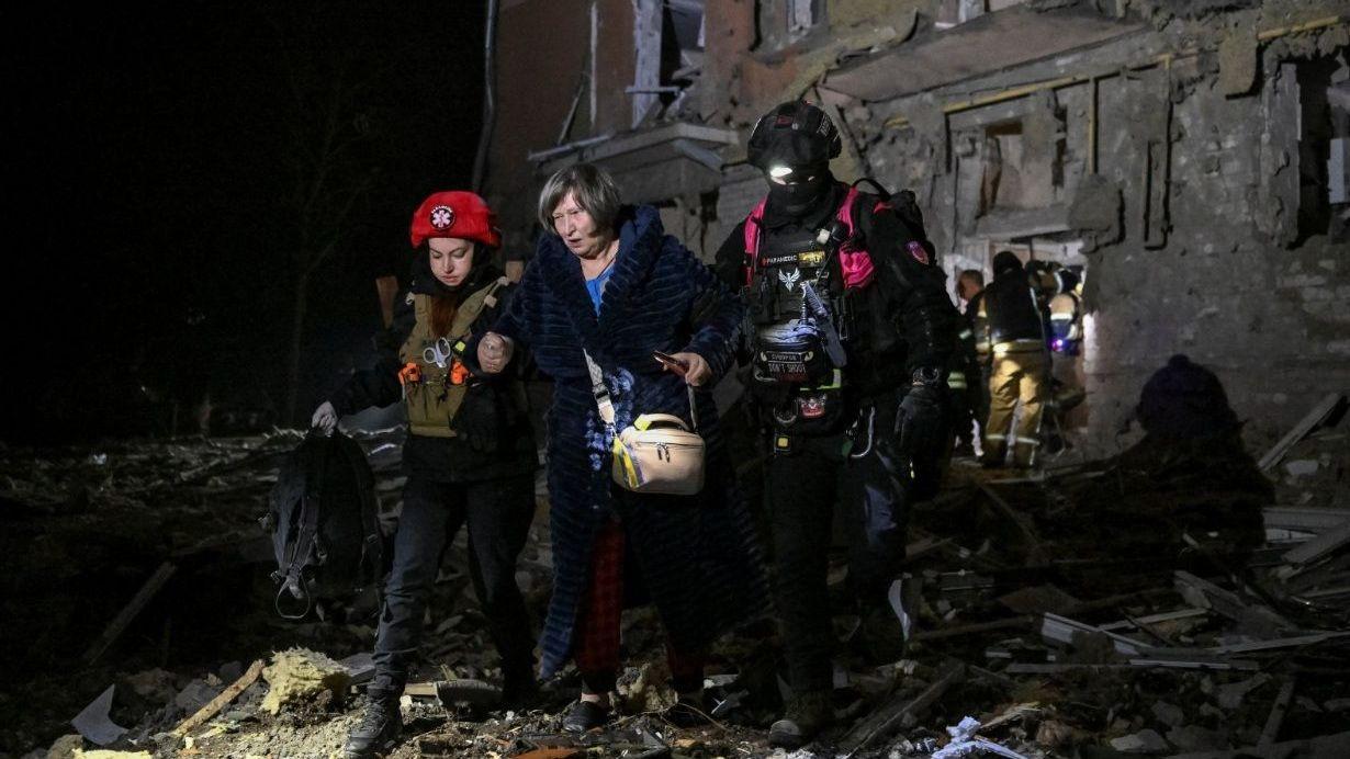 Plano general que muestra a una mujer de mediana edad alejándose de un edificio residencial dañado por ataques rusos durante la noche. Va de la mano de dos rescatistas que la ayudan a caminar sobre los escombros.