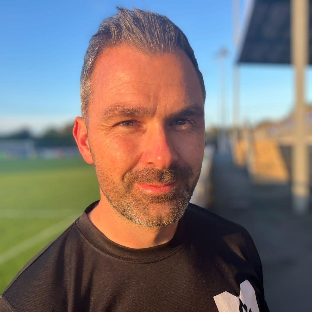 Primer plano de Gavin Roberts de pie junto a un campo de fútbol bajo la cálida luz del atardecer. Gavin lleva una camiseta deportiva negra, con el fondo ligeramente desenfocado para mostrar parte del campo a la izquierda. Tiene el pelo corto y gris con flequillo de punta. También luce una barba corta y gris. 