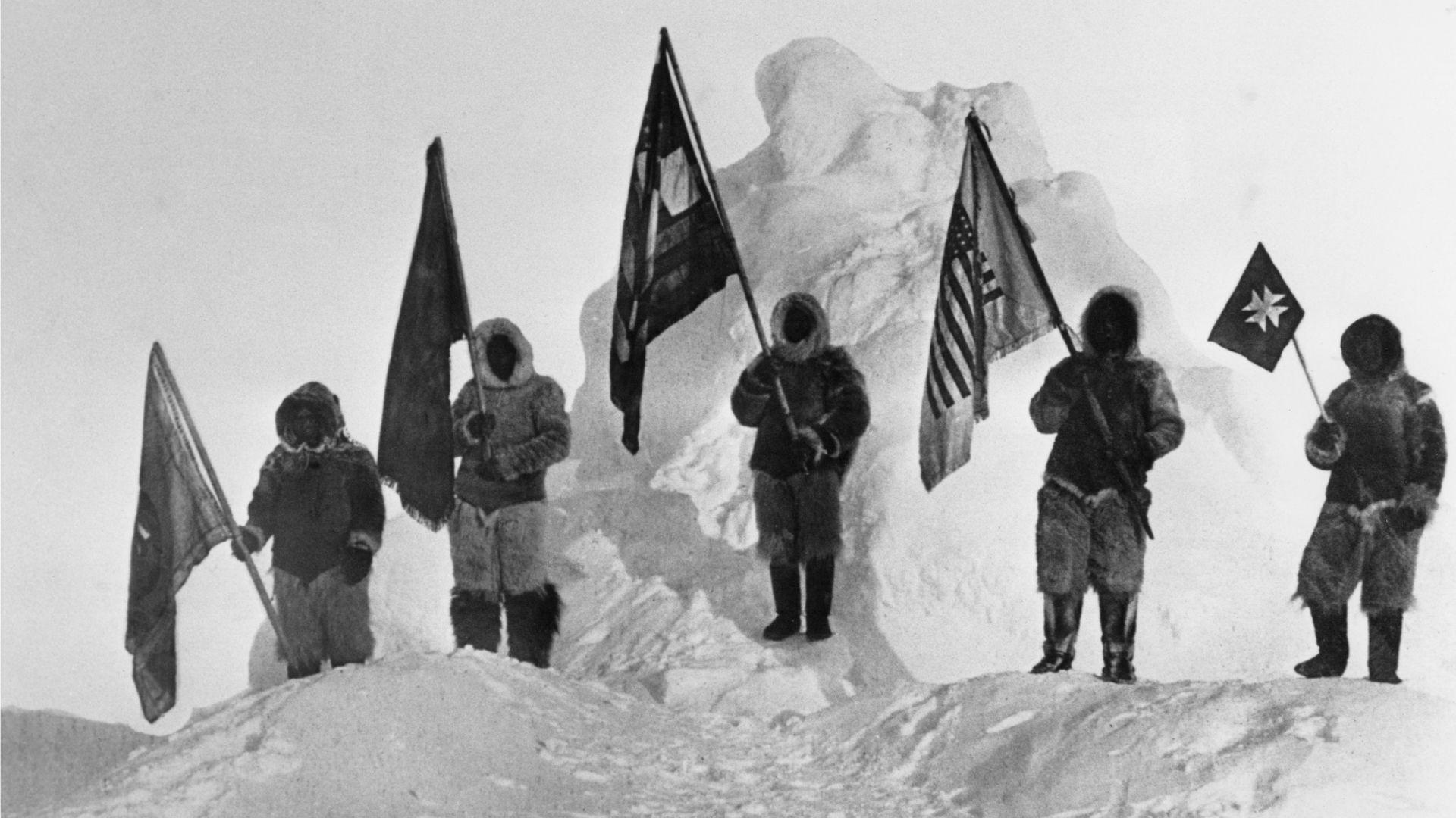 Una fotografía en blanco y negro de cinco personas vestidas con ropa gruesa, cada una sosteniendo una gran bandera, de pie sobre un suelo cubierto de nieve frente a un montón de nieve.