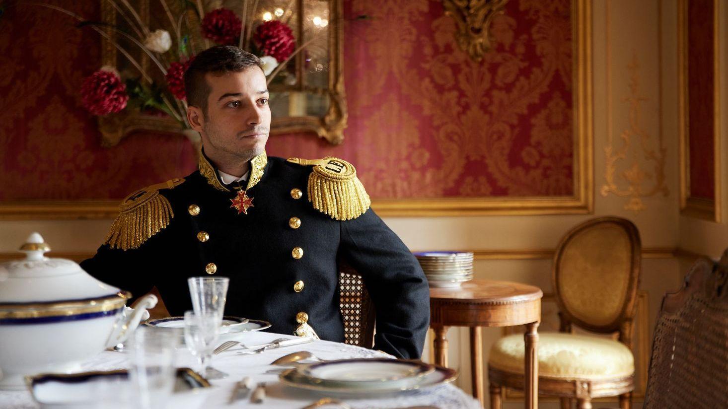 Jonas Lauwiner, sentado a la mesa y vestido con un uniforme militar, en uno de los salones de su "palacio". Sobre la mesa hay platos y porcelana fina, mientras las paredes están cubiertas de alfombras rojas y doradas.
