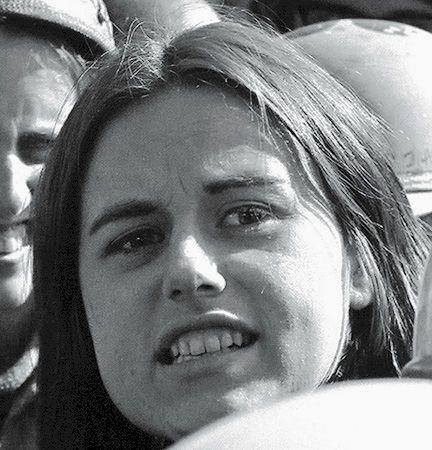 Primer plano del rostro de una joven de cabello castaño, con otras personas detrás. Tiene los labios entreabiertos, como si estuviera a punto de gritar. Está mirando a alguien o algo que se encuentra detrás de la cámara.