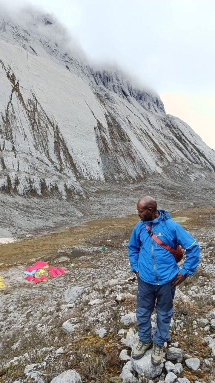 Justinus, un hombre de la tribu Moni, está de pie frente a la montaña. Lleva una chaqueta azul y pantalones azul oscuro, con una mochila roja cruzada en el pecho