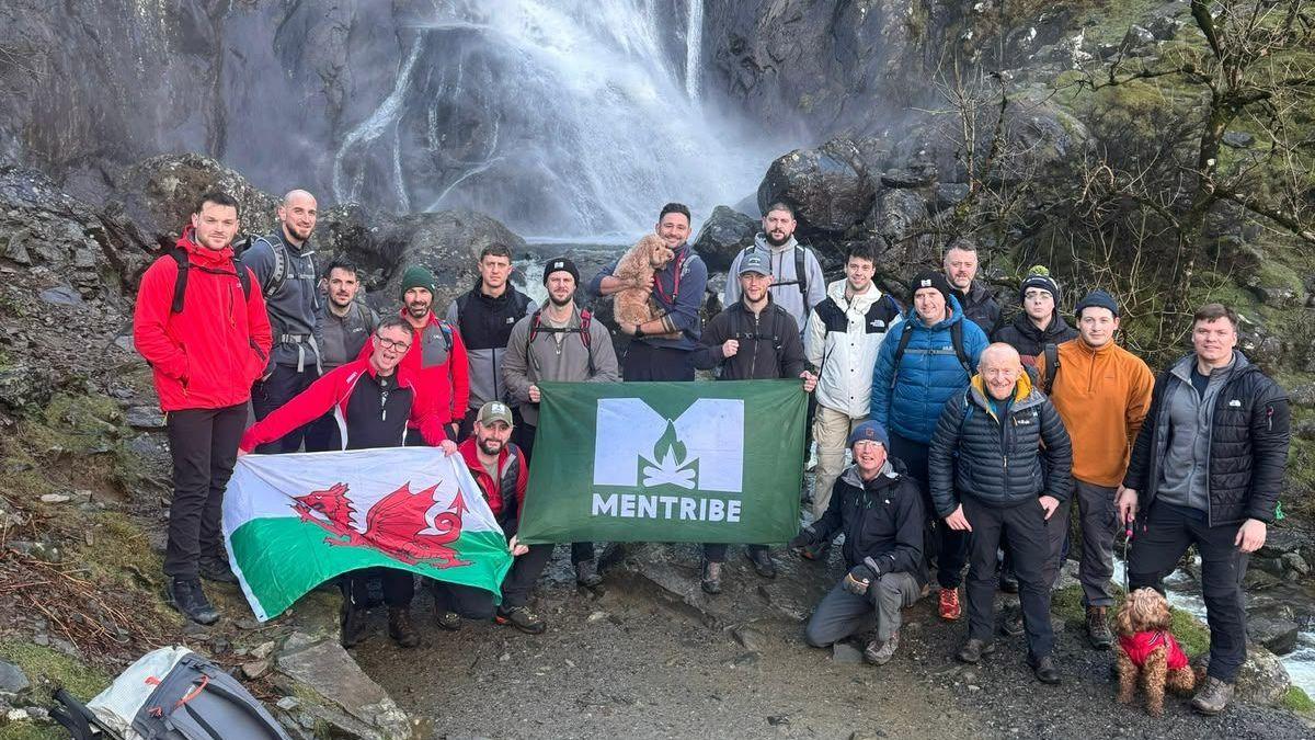 Un numeroso grupo de excursionistas posa al pie de una cascada que cae por un escarpado acantilado rocoso. El grupo se encuentra en un sendero rocoso rodeado de rocas cubiertas de musgo y árboles desnudos. Varias personas portan una bandera galesa y una pancarta verde con la inscripción "MENTRIBE". Se aprecian dos perros: uno en brazos de un participante y otro con un abrigo rojo cerca del frente. Las mochilas reposan sobre las rocas en primer plano, mientras que la bruma de la cascada se eleva tras el grupo en un paisaje montañoso al aire libre.