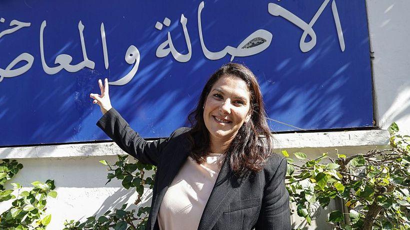 El Mansouri, vestida con una chaqueta negra sobre una camisa blanca, posa frente al lema de su partido, escrito en árabe con letras blancas sobre un fondo azul.