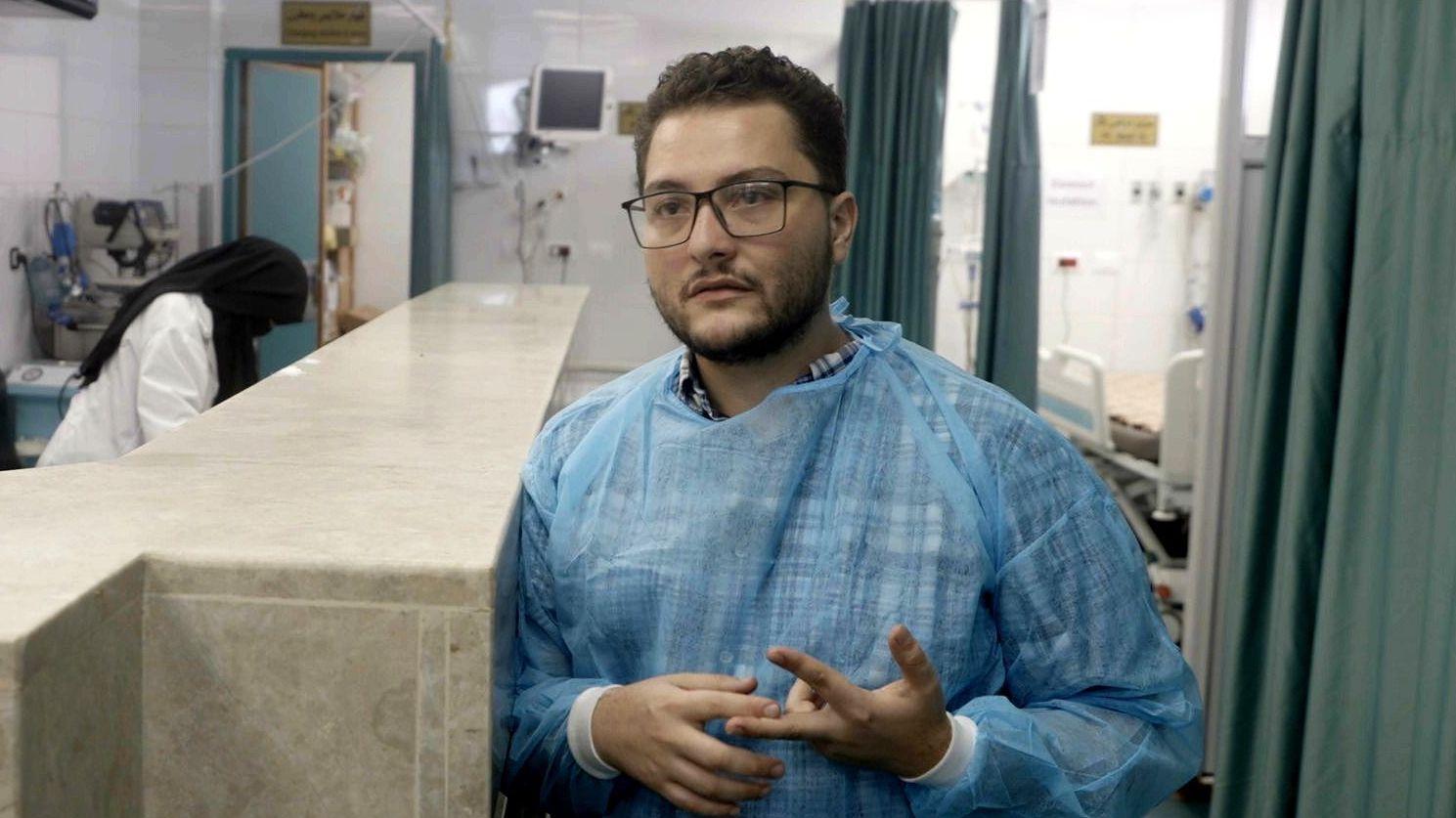 En la fotografía, el doctor Awni al-Jaafarawi aparece vestido con una bata médica azul en una sala de hospital. Detrás de él se puede ver a una mujer con un pañuelo en la cabeza.