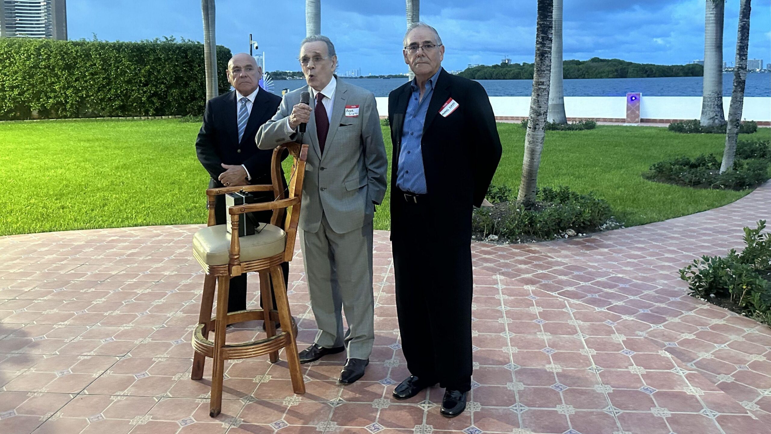 Juan Omar Sixto habla por un micrófono, de pie entre dos de sus compañeros. Están de pie frente a un taburete sobre el que hay un altavoz.