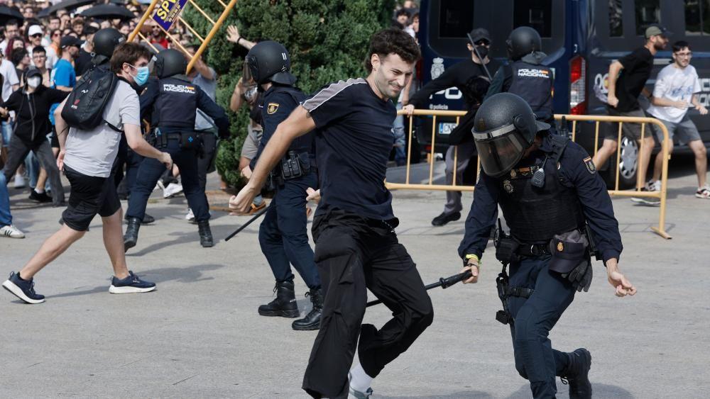 Un agente antidisturbios parece sostener una porra cerca de las piernas de un manifestante. Al fondo se observa una multitud de manifestantes y varios agentes antidisturbios uniformados con cascos. Al fondo, se ve a alguien levantando parte de una valla amarilla.