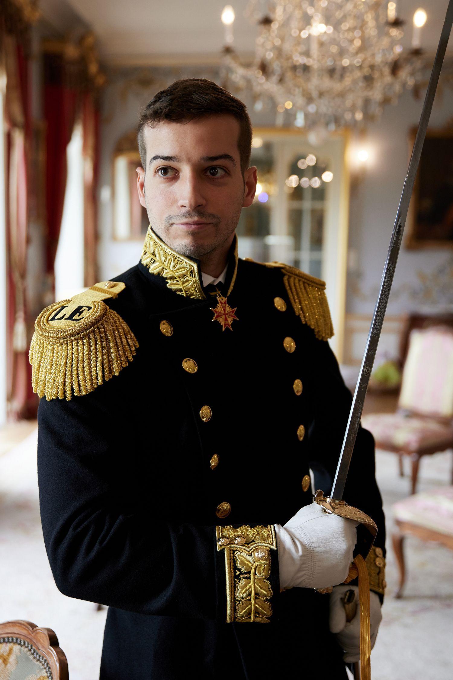 Jonas Lauwiner viste su tradicional uniforme negro con bordes dorados y lleva una espada. Al fondo se ve una de las salas de su "palacio", con grandes ventanales, sillas y candelabros.