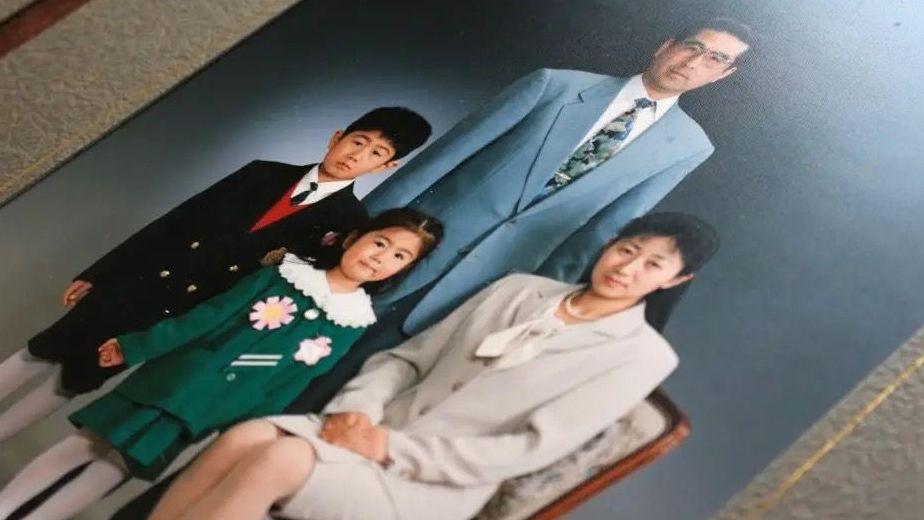 Un retrato familiar formal muestra a cuatro personas posando frente a un fondo oscuro de estudio: una mujer sentada en una silla con un traje claro en primer plano, un hombre con un traje azul claro de pie detrás, y dos niños a sus lados. El niño más pequeño lleva un vestido verde con grandes adornos florales y el mayor viste un traje oscuro con chaleco rojo y calcetines blancos. Él tiene expresión seria, mientras que los demás sonríen levemente.