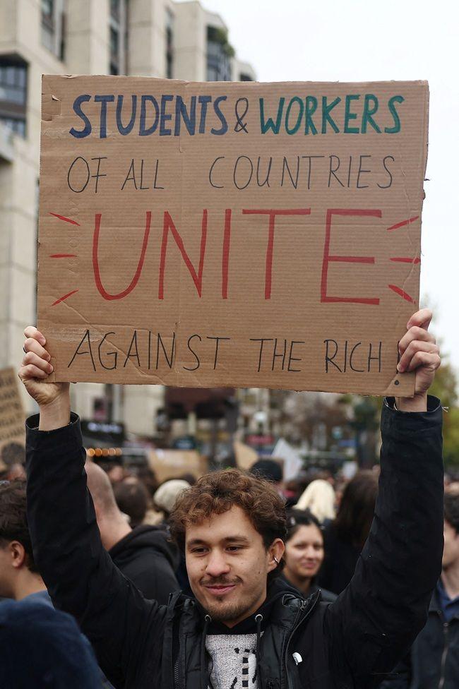 Un joven, con una chaqueta negra y en medio de una concentración de personas, levanta un cartel que dice: "Students & workers of all countries unite against the rich".

