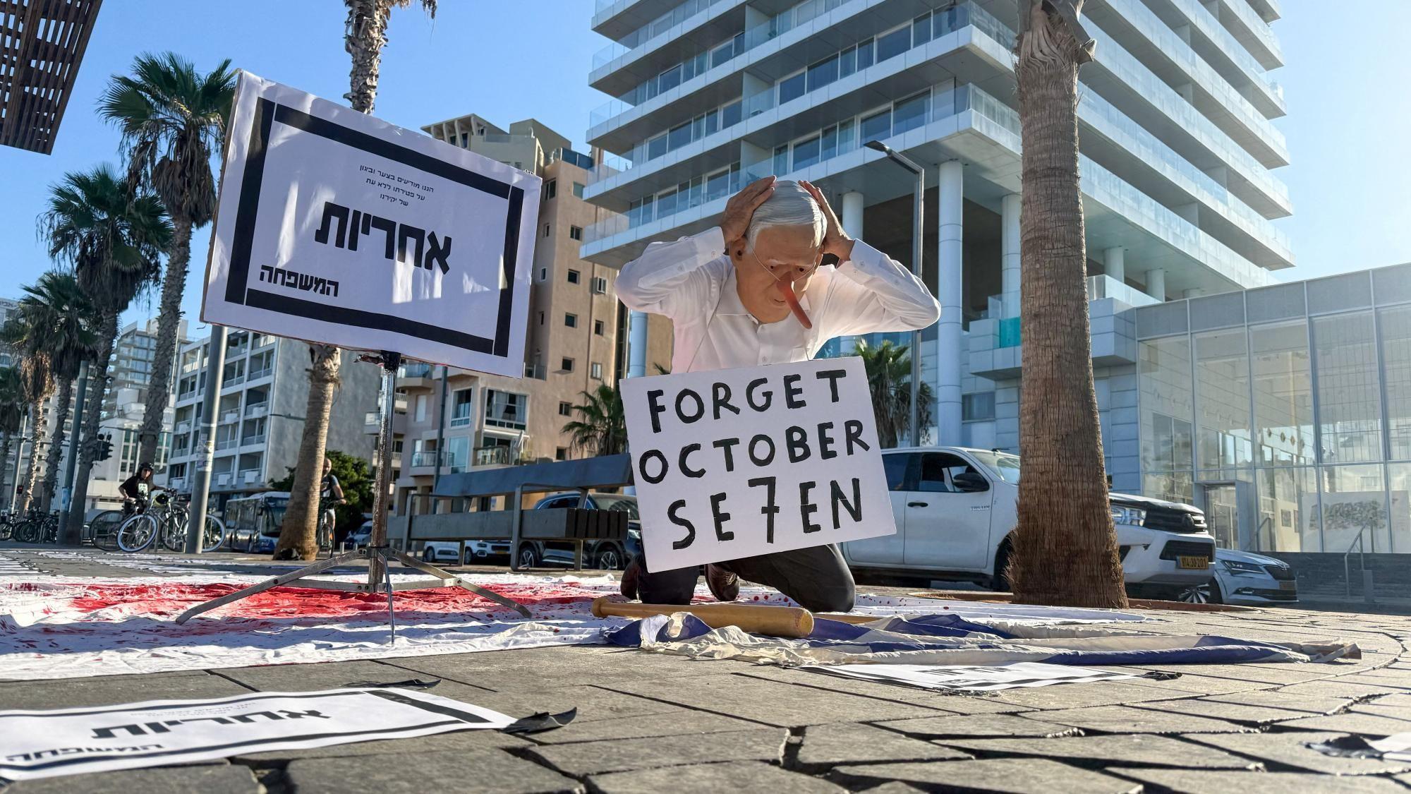 Un manifestante con una mascarilla que muestra al primer ministro israelí Benjamin Netanyahu se arrodilla sobre una sábana cubierta de pintura roja con un cartel en hebreo que anuncia la 