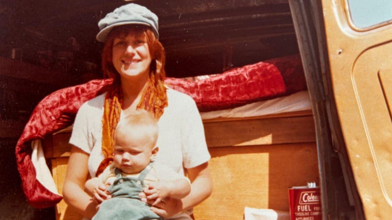 Una joven con gorra clara y bufanda naranja está sentada en la puerta abierta de una furgoneta, con un niño pequeño vestido con un mono pálido en su regazo. Dentro de la furgoneta, detrás de ellos, hay una cama cubierta con una colcha roja. Cerca de la puerta del vehículo se encuentra un bidón de combustible rojo con la inscripción "Coleman".