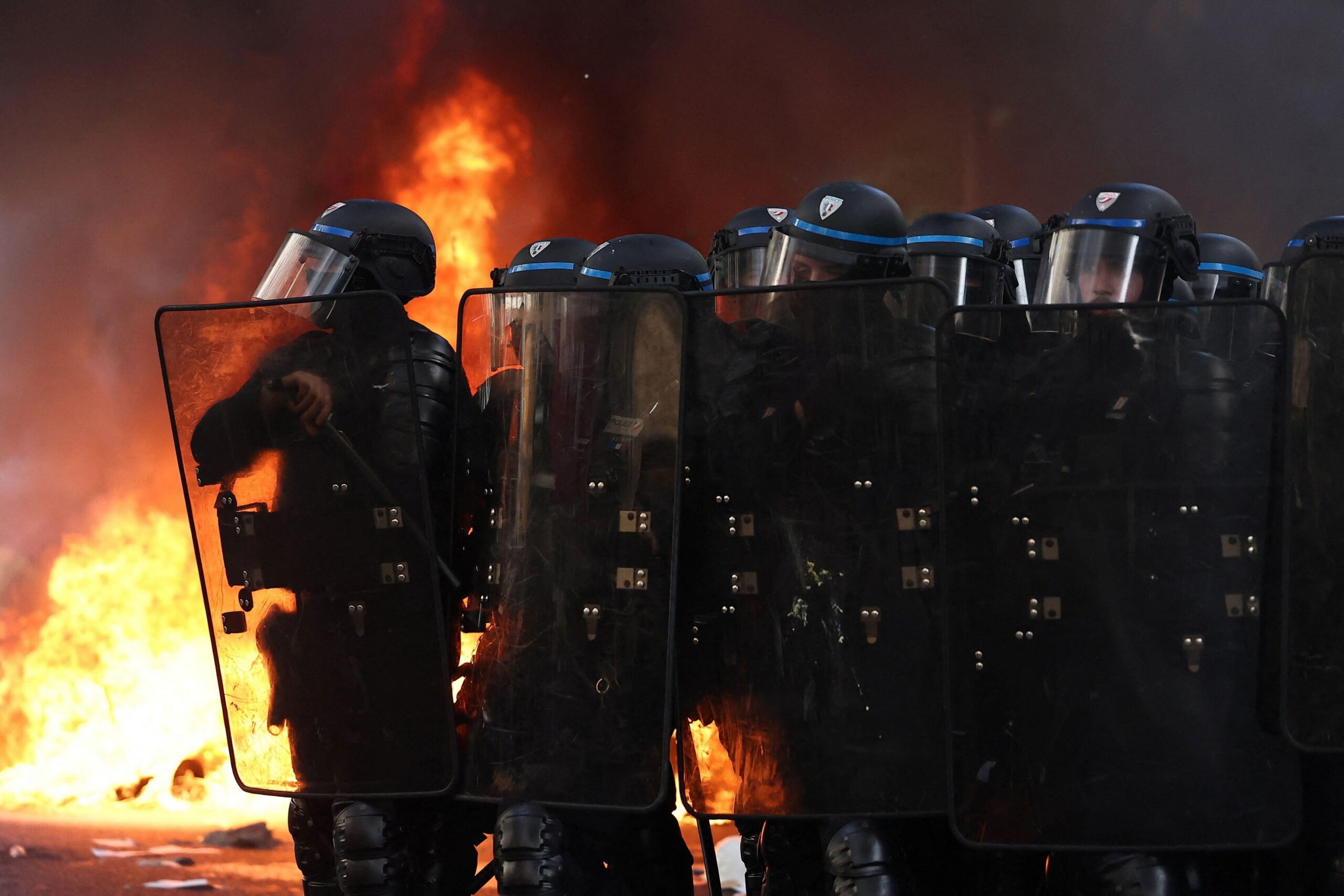 Un grupo de policía antimotines, con escudos, cascos y macanas, al frente de un incendio en la calle.