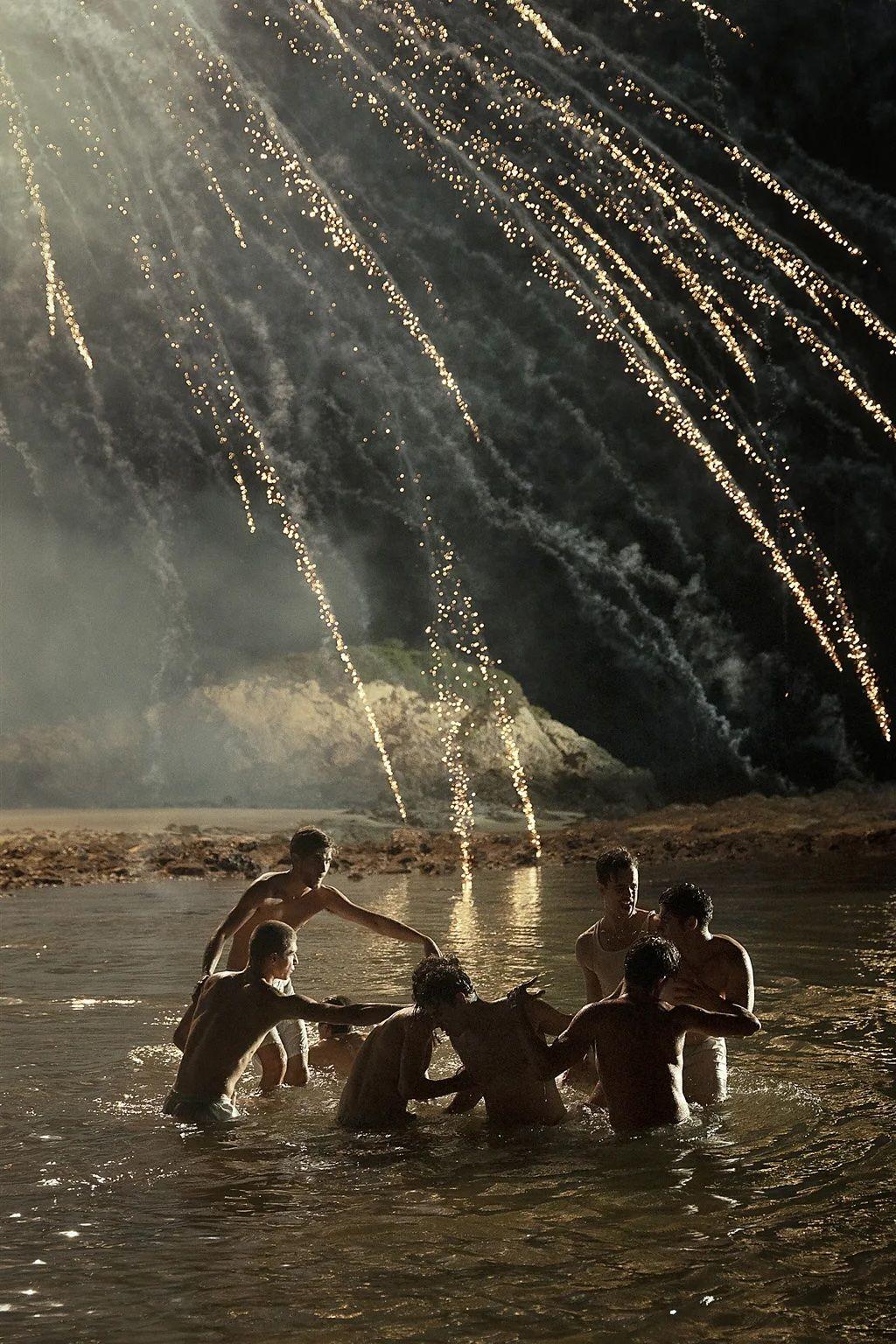 Un grupo de hombres sin camiseta en un lago de noche, mientras estallan fuegos artificiales detrás suyo.