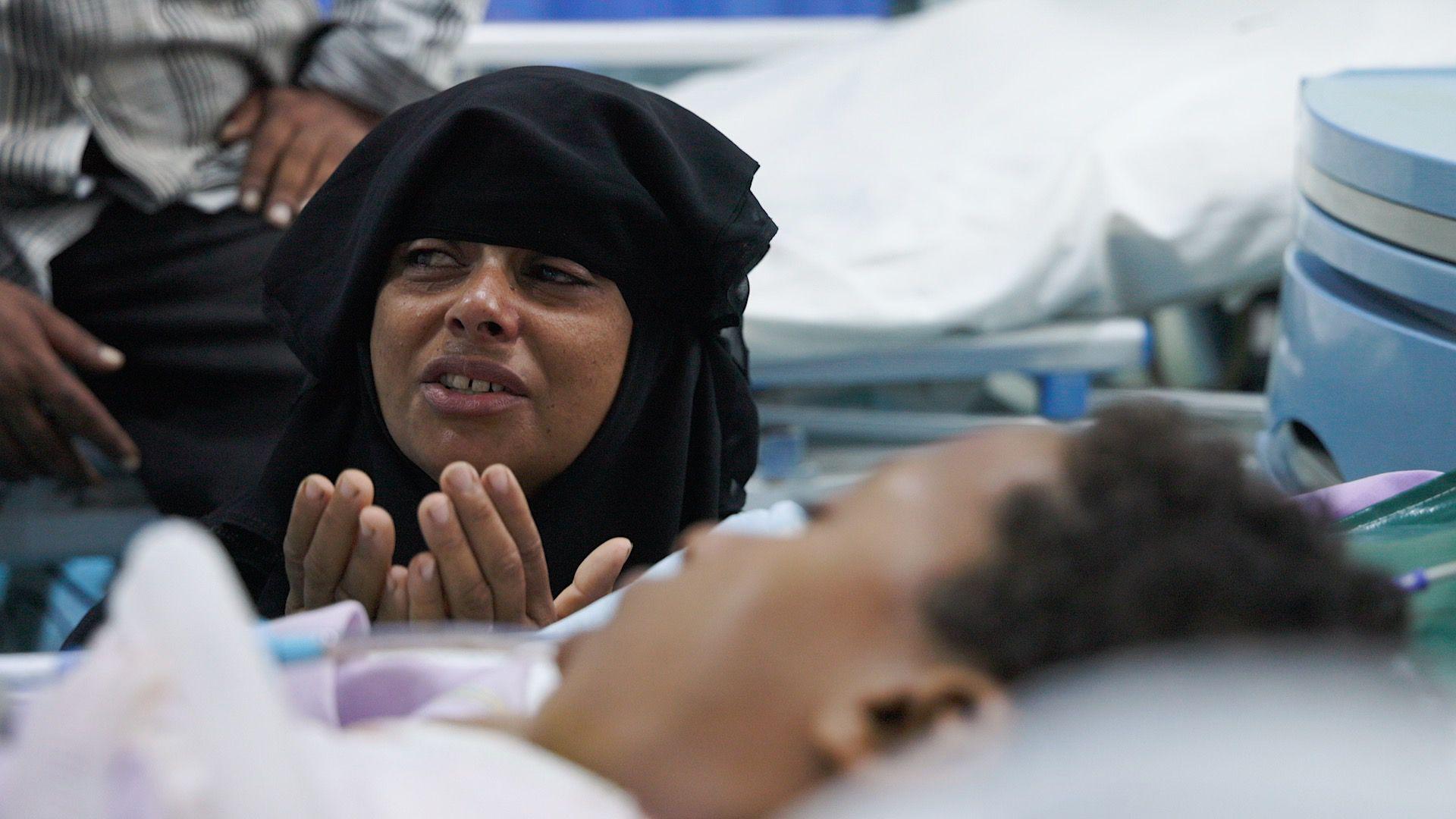 Una mujer angustiada, con un pañuelo negro en la cabeza, está sentada junto a una cama de hospital, con las manos levantadas hasta la barbilla. Un niño yace en la cama frente a ella.