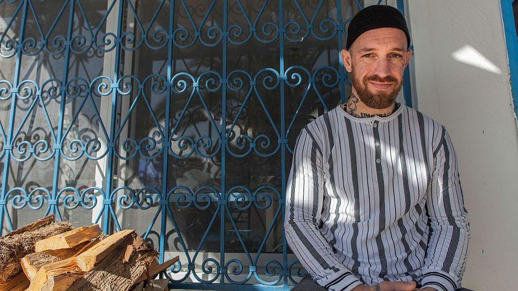 "Fergie" Chambers, sonríe sentado junto a una ventana con una reja azul.  A su derecha tiene unos pedazos de leña. 