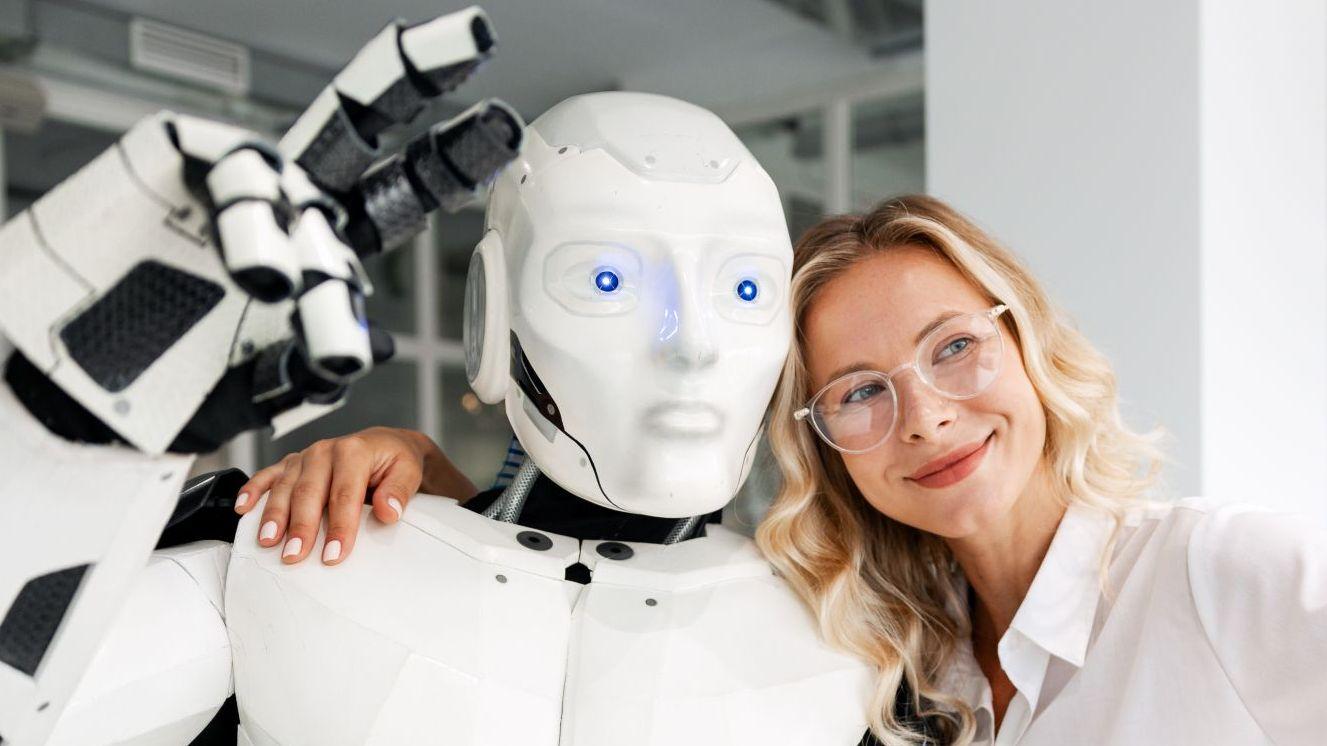 Una mujer de pelo rubio y con anteojos, sonríe mientras abraza a un robot de ojos azules que hace la señala de paz con dos dedos 