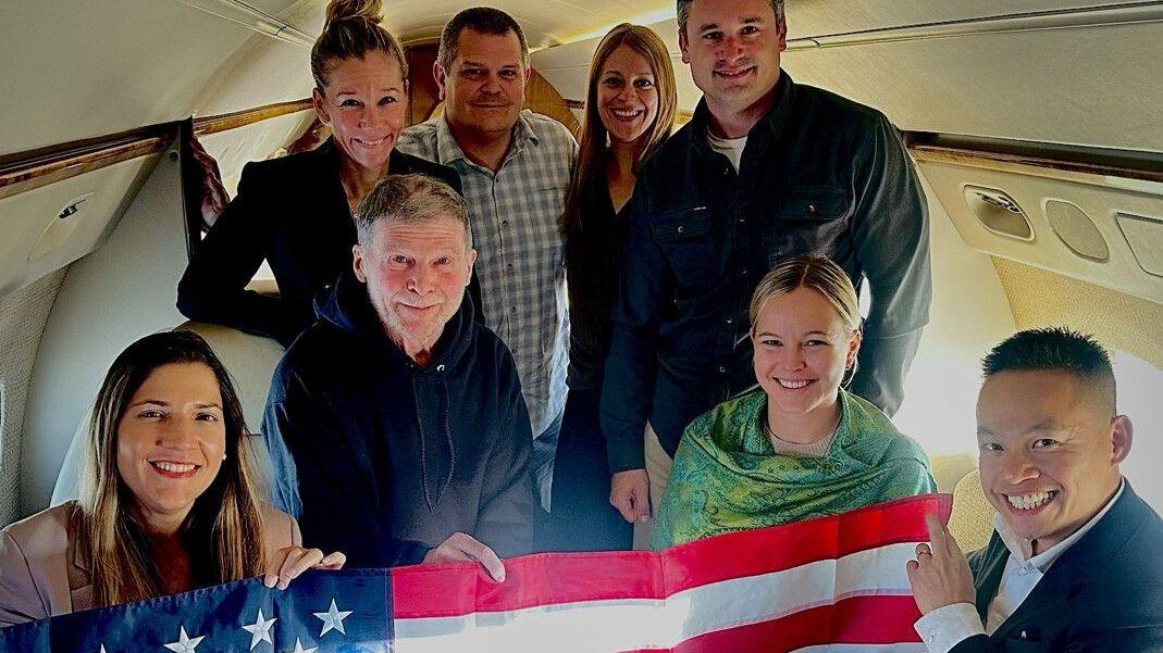 Stephen Hubbard (segundo por la izquierda, sentado), fotografiado con miembros de su familia sosteniendo una bandera estadounidense dentro de un avión. En la foto aparecen ocho personas: cuatro de pie y cuatro sentadas.