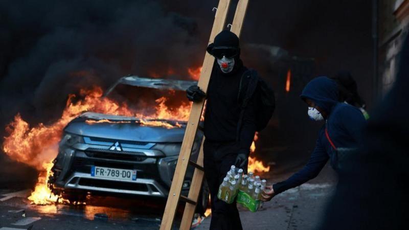 Protestas en Francia. Se ve un auto en llamas y varios hombres enmascarados. 