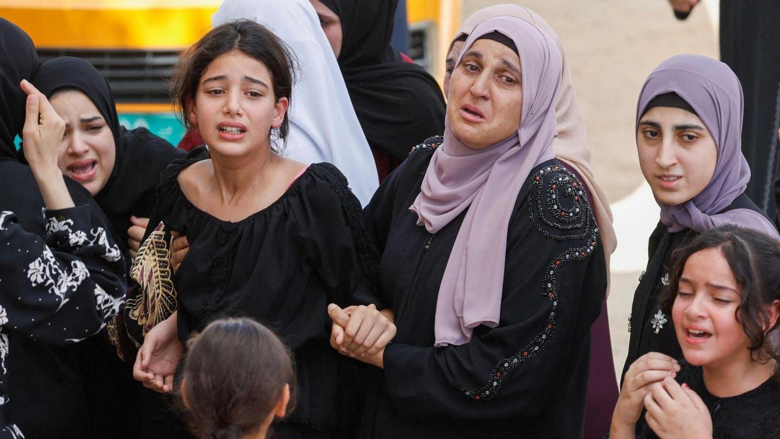 Una mujer vestida con un vestido negro y un pañuelo morado en la cabeza sostiene la mano de una niña con una blusa negra, como en un funeral. Se ve a otras tres niñas vestidas de negro; una lleva un pañuelo morado y la otra, uno negro. Todas lloran.