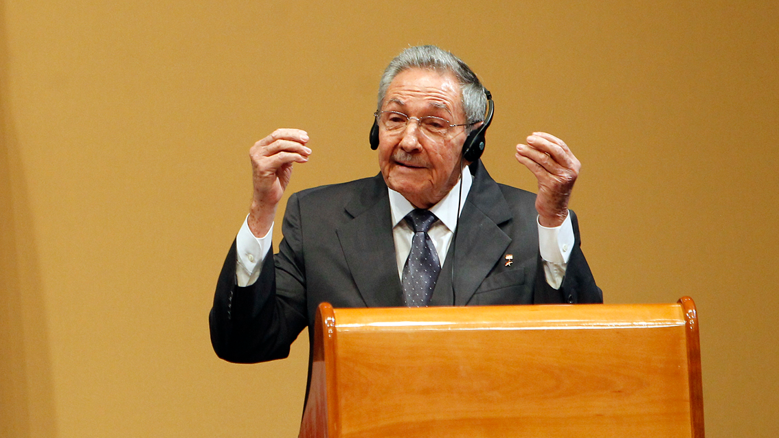Raúl Castro, vestido con una corbata y un traje grises y una camisa blanca, habla eun un podio de madera. Lleva unos audífonos y lentes