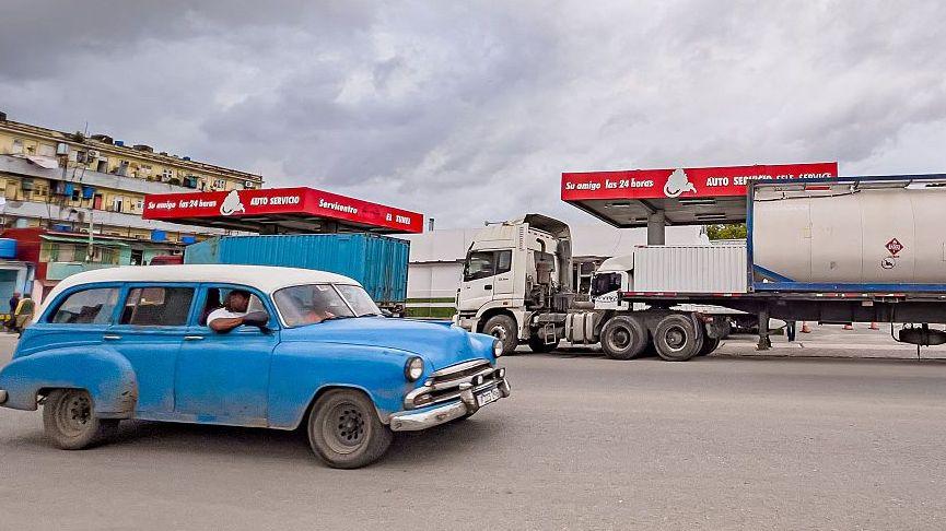 Un automóvil estadounidense antiguo pasa junto a un camión de una empresa privada cubana (mipyme) que transporta un isotanque de combustible importado y está estacionado frente a una gasolinera.